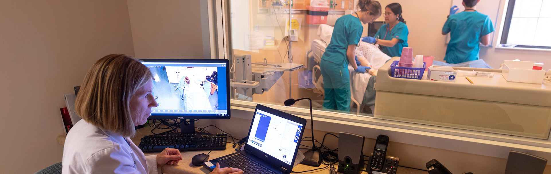 A female School of Nursing faculty looks at a laptop and larger screen. She is observing three nursing students through a large window in the simulation learning center as they perform tasks on a manikin. 