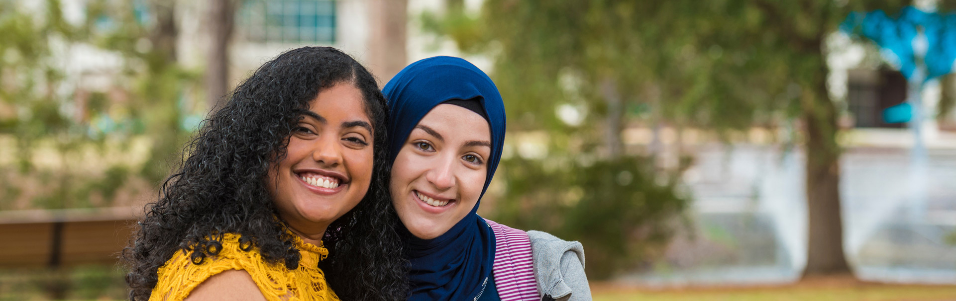 Two students sitting on a bench outside, smiling at the camera. 