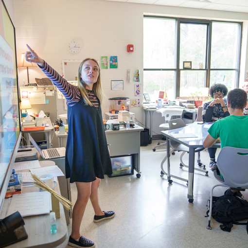 Teacher stands at front of class pointing at the white board