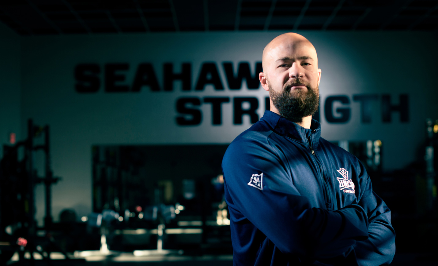 Bradley Moore, the director of Athletics Department Sports Performance Office, stands with his arms crossed in a UNCW athletics shirt in the student recreation center.