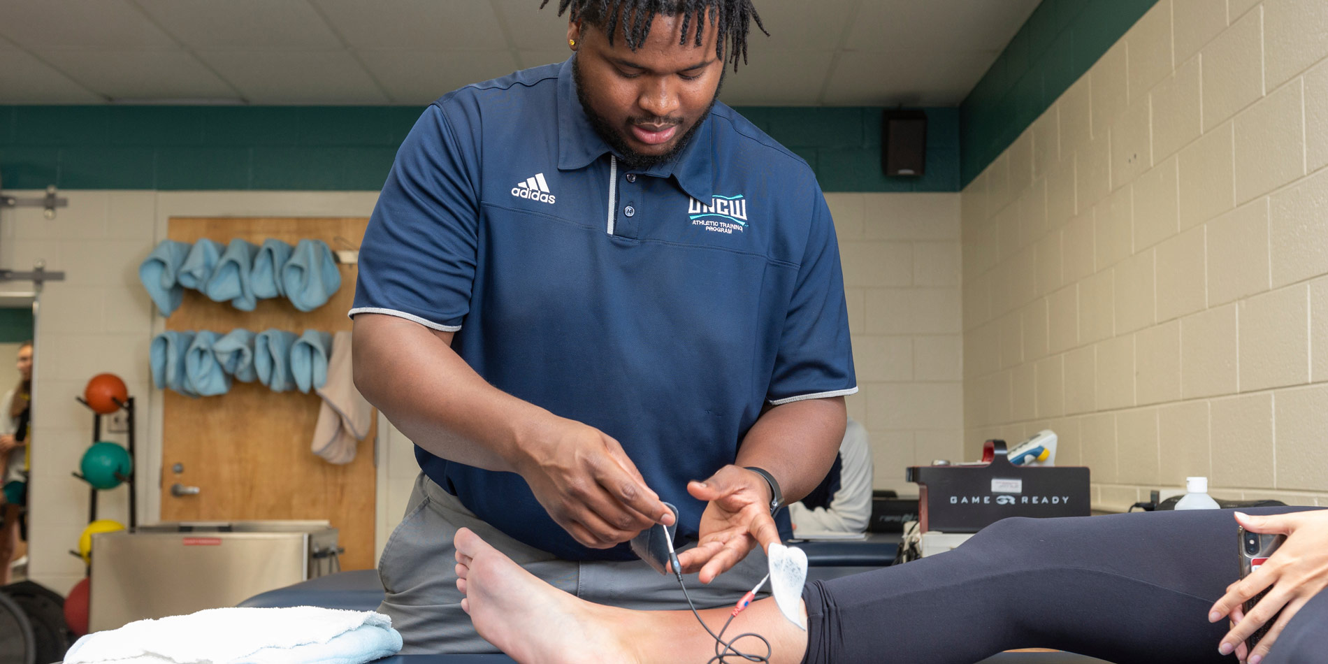 A group of students simulate a situation in which they will use their knowledge as Athletic Trainers. In the picture, two students assist another to a bed where a nurse is waiting to help.