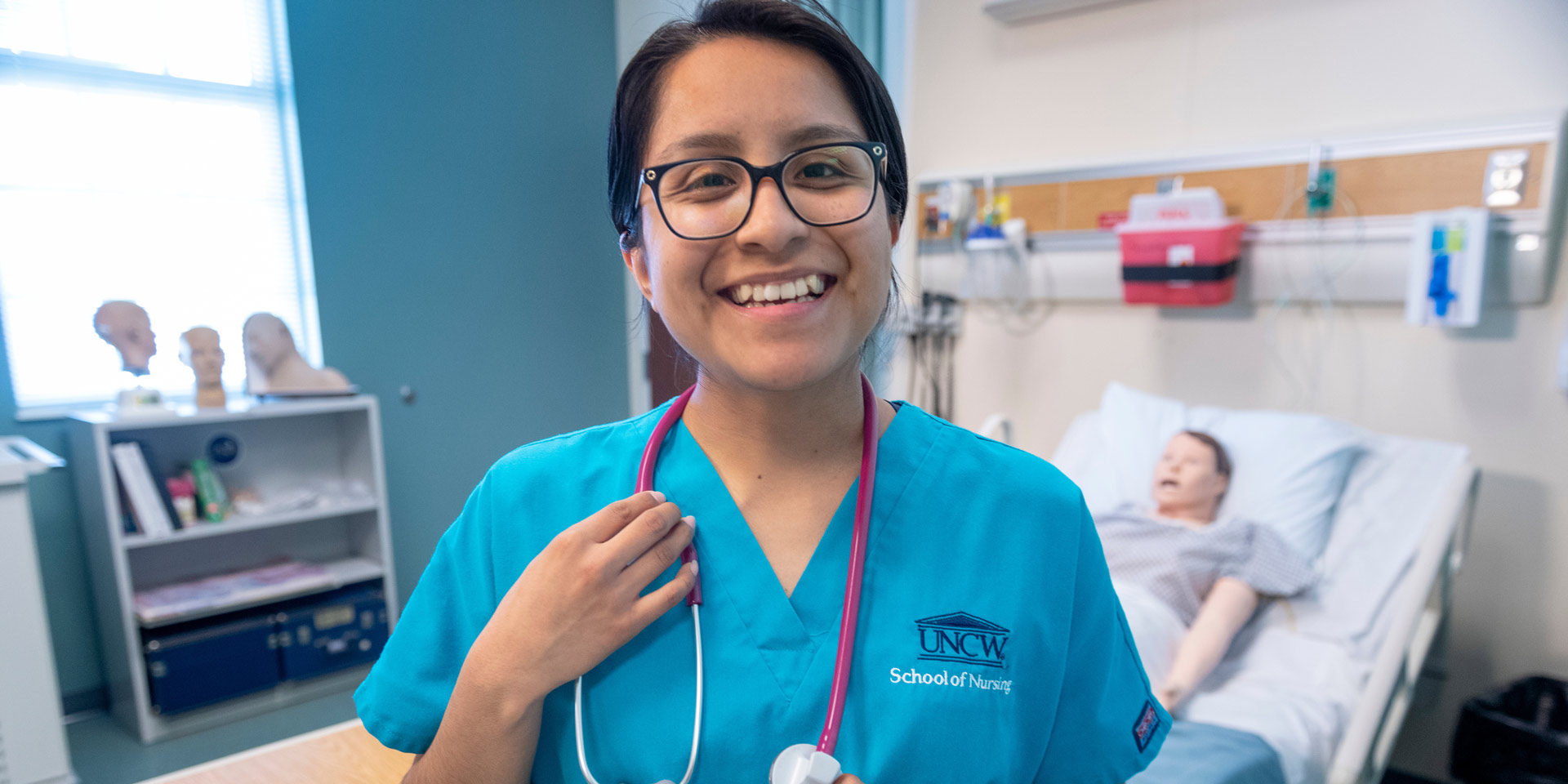 A student in School of Nursing scrubs smiles for the camera in a mock patient room