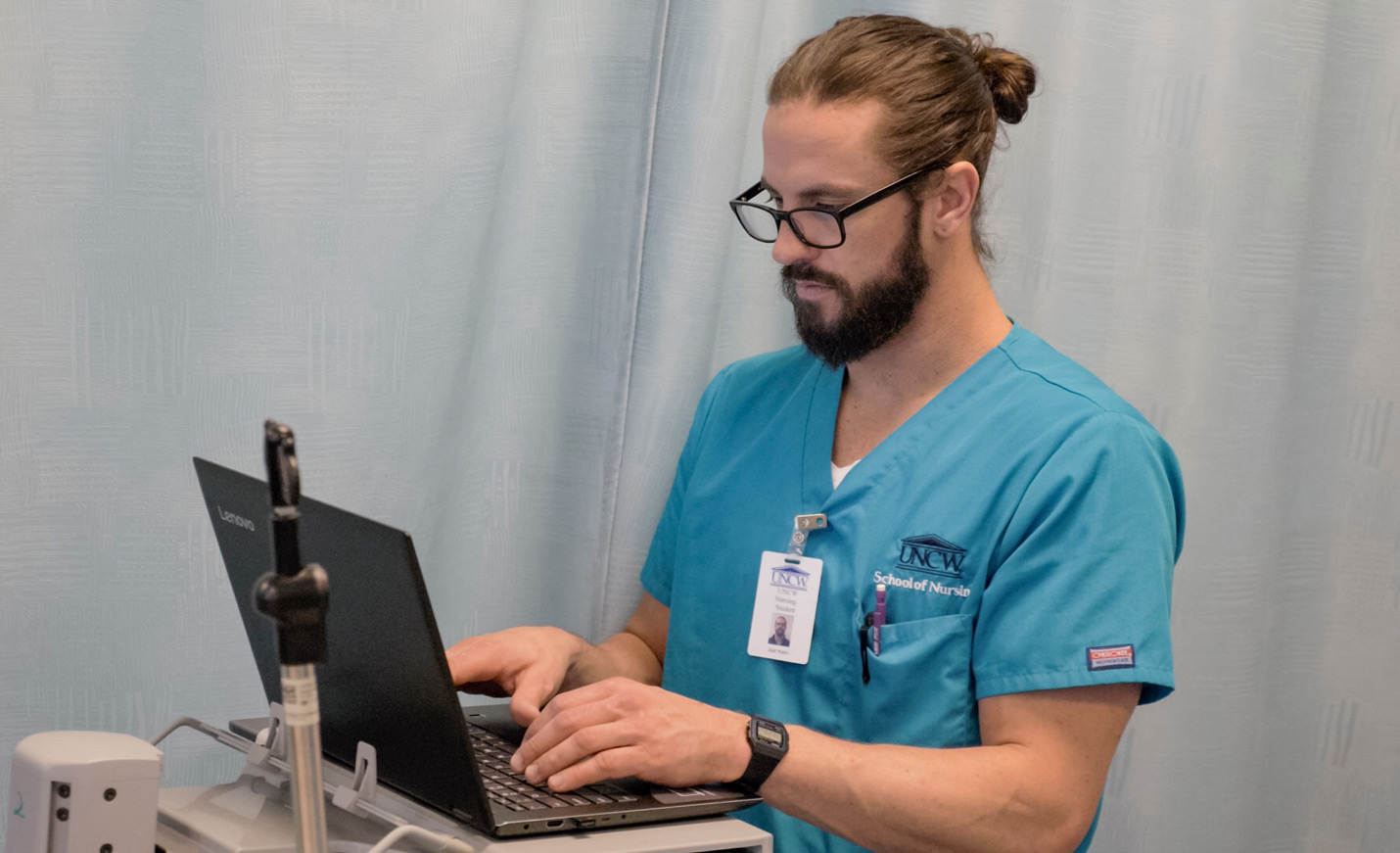 A nursing students, wearing scrubs, types on the keyboard of his laptop.