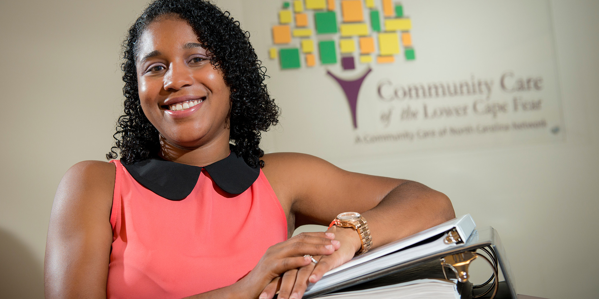 An employee of Community Care of the Lower Cape Fear smiles for the camera in front of a logo on the wall