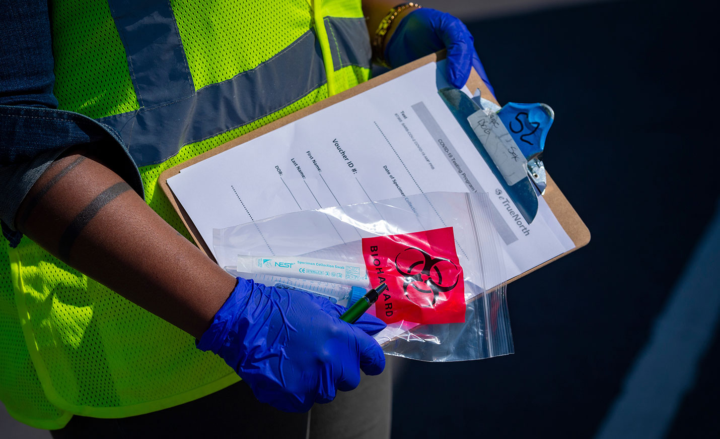 Someone in a safety vest wearing gloves holds a clipboard and a biohazard bag.