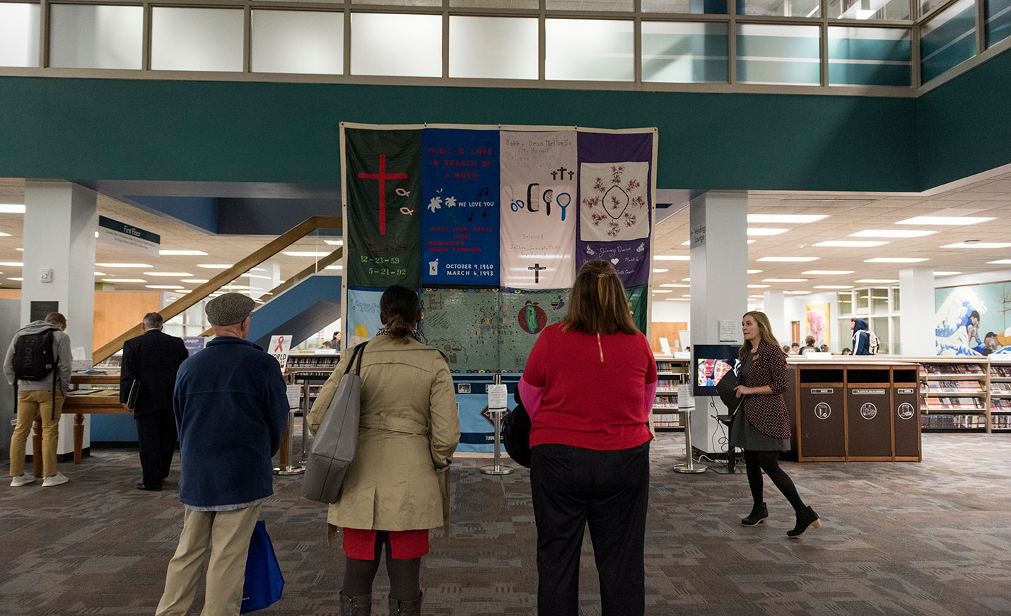 Three people stand with their backs to the camera as they look up at the AIDS quilt in UNCW Library.