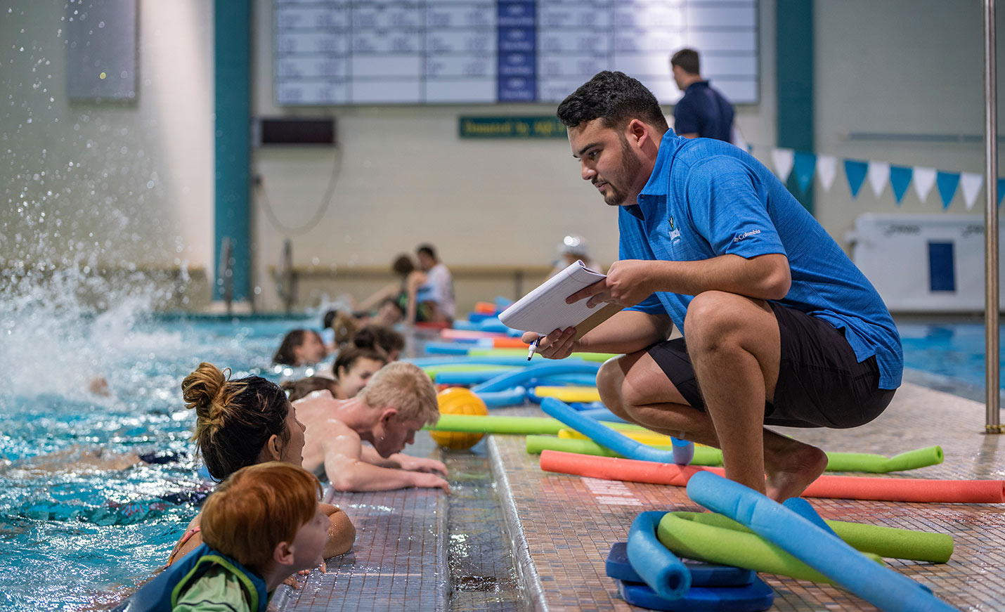 A large swim class hangs onto the edge of the pool while their instructor squats next to the edge with a clipboard.