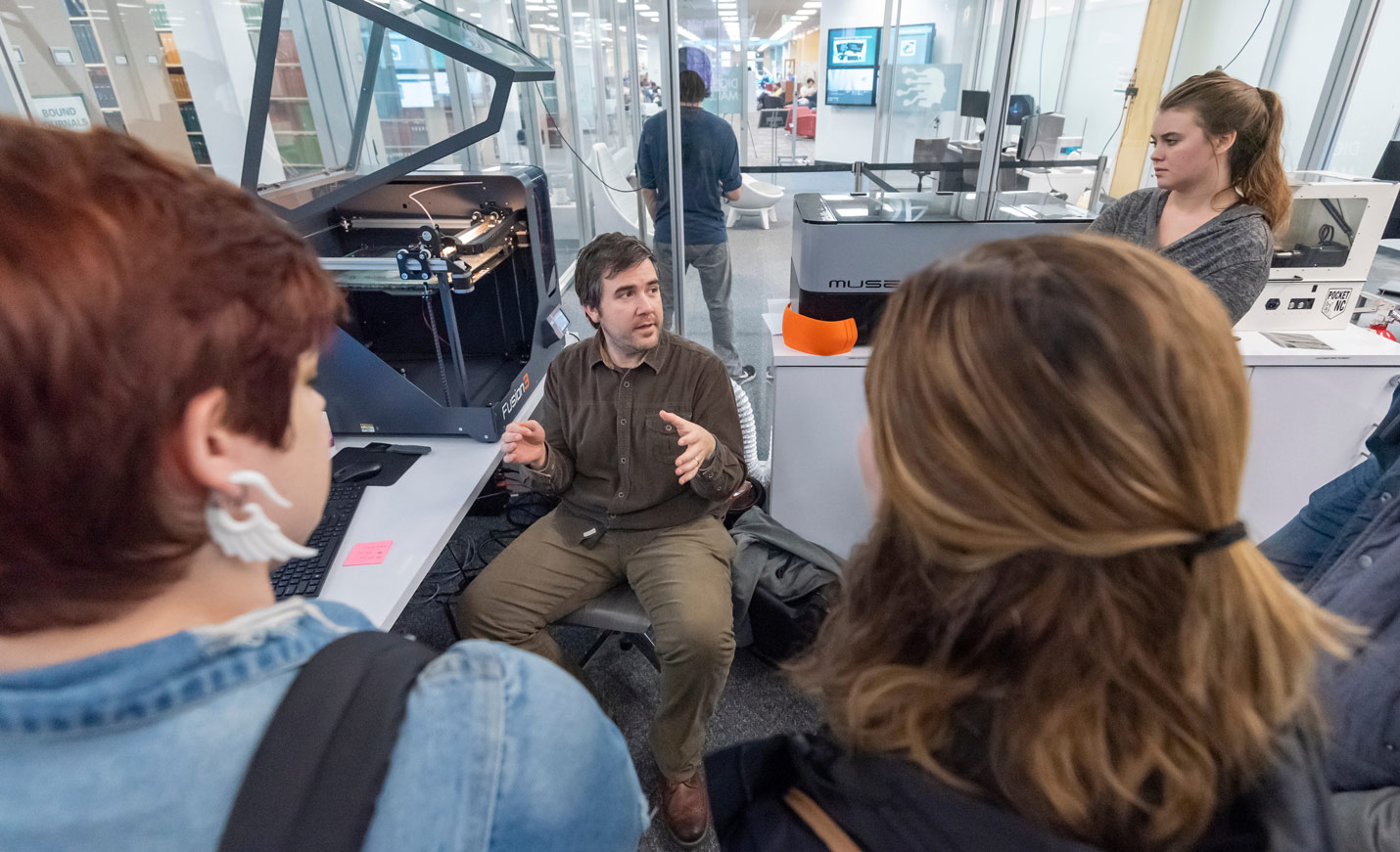 Three students listen to an instructor, who is seated in front of a 3-D Printer