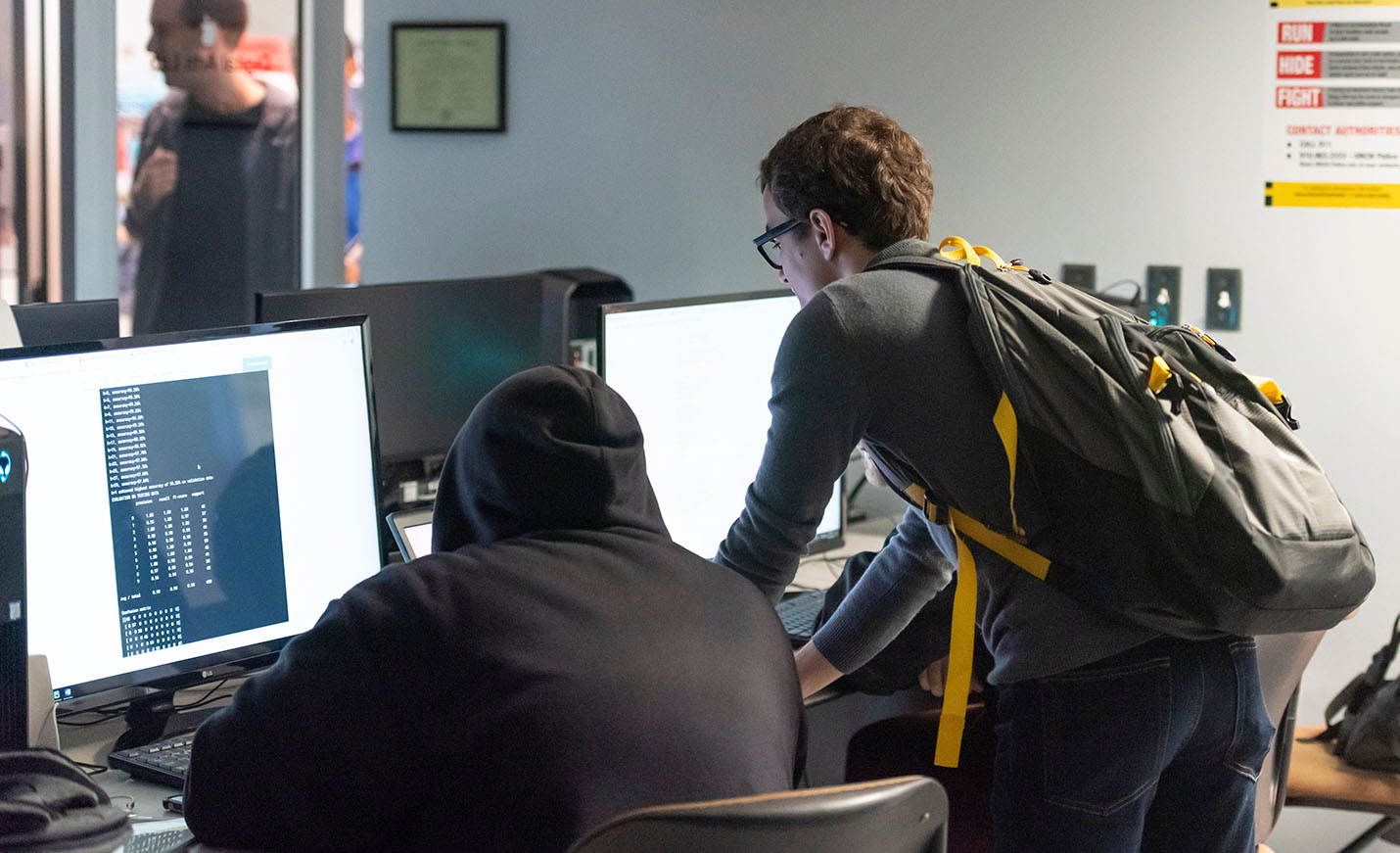 Two students - one seated, one standing - work in front of computers