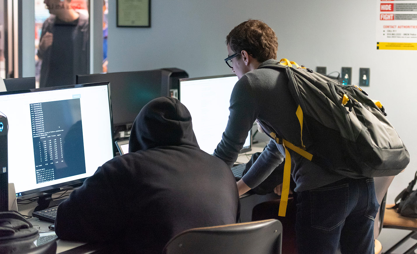 Two students - one seated, one standing - work in front of computers