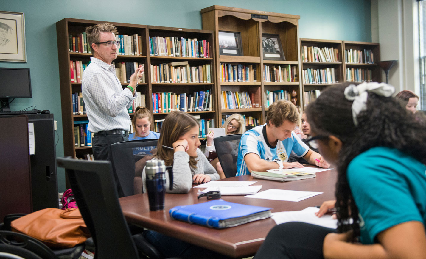 An instructor standing in front of several shelves of books addresses students who are seated at a table