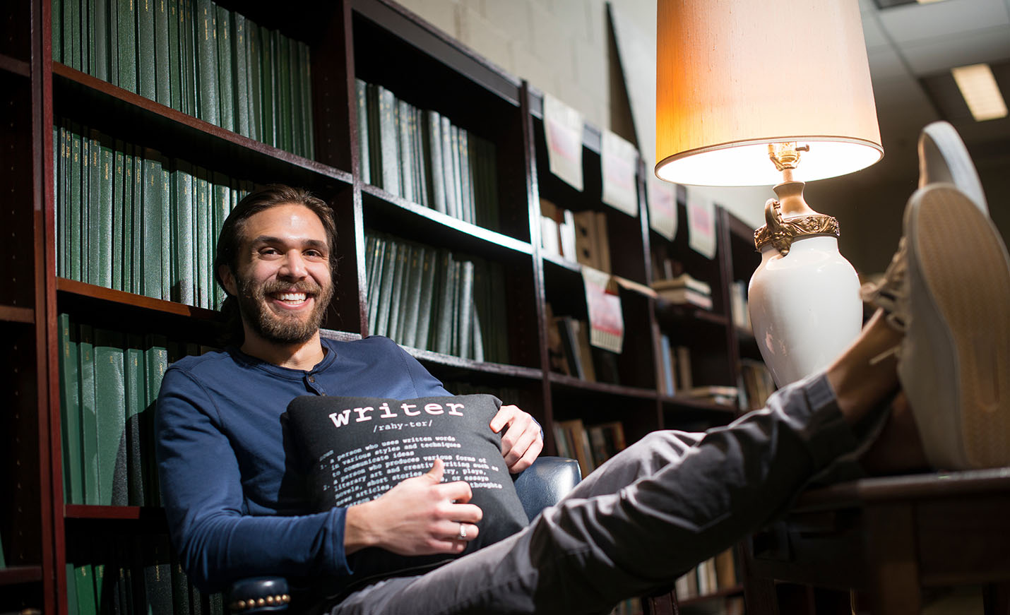 creative writing student sitting in library holding a pillow with the word writer