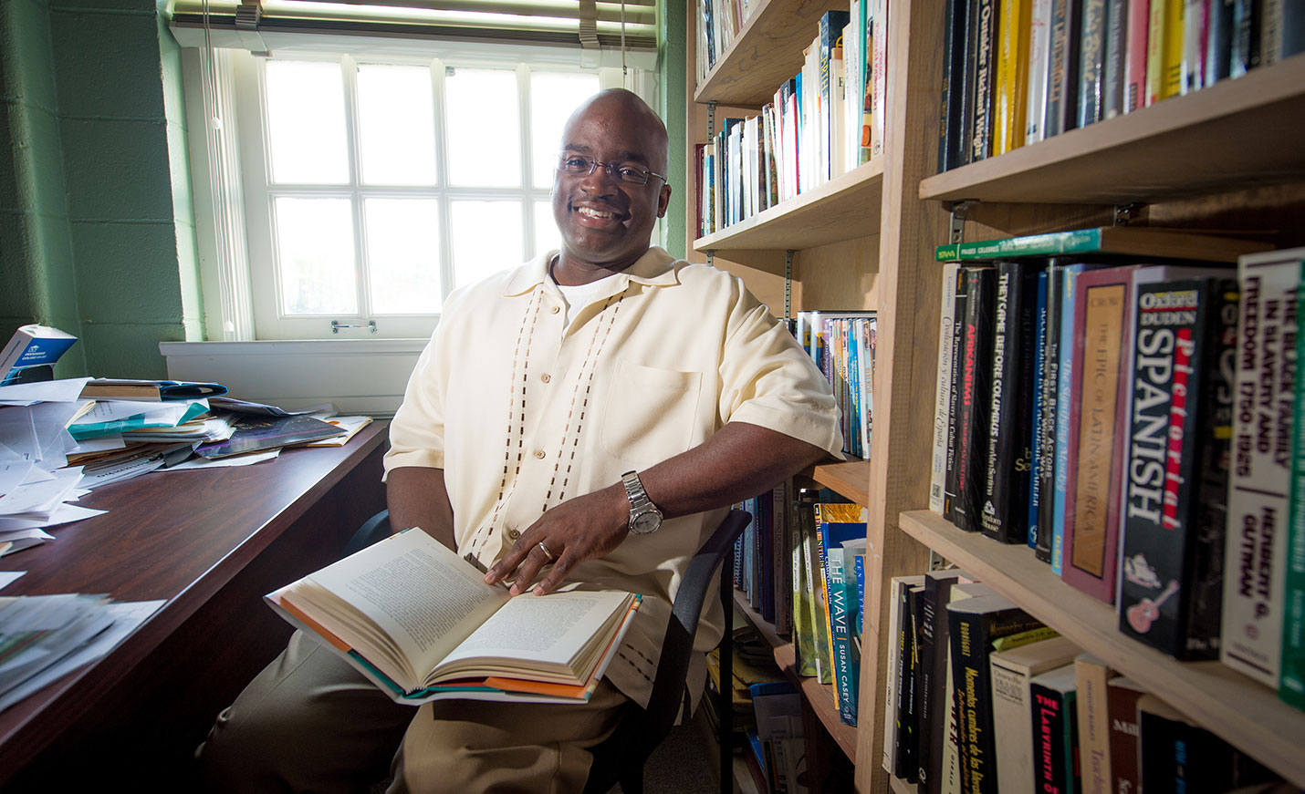 faculty member in office with book open on his lap.