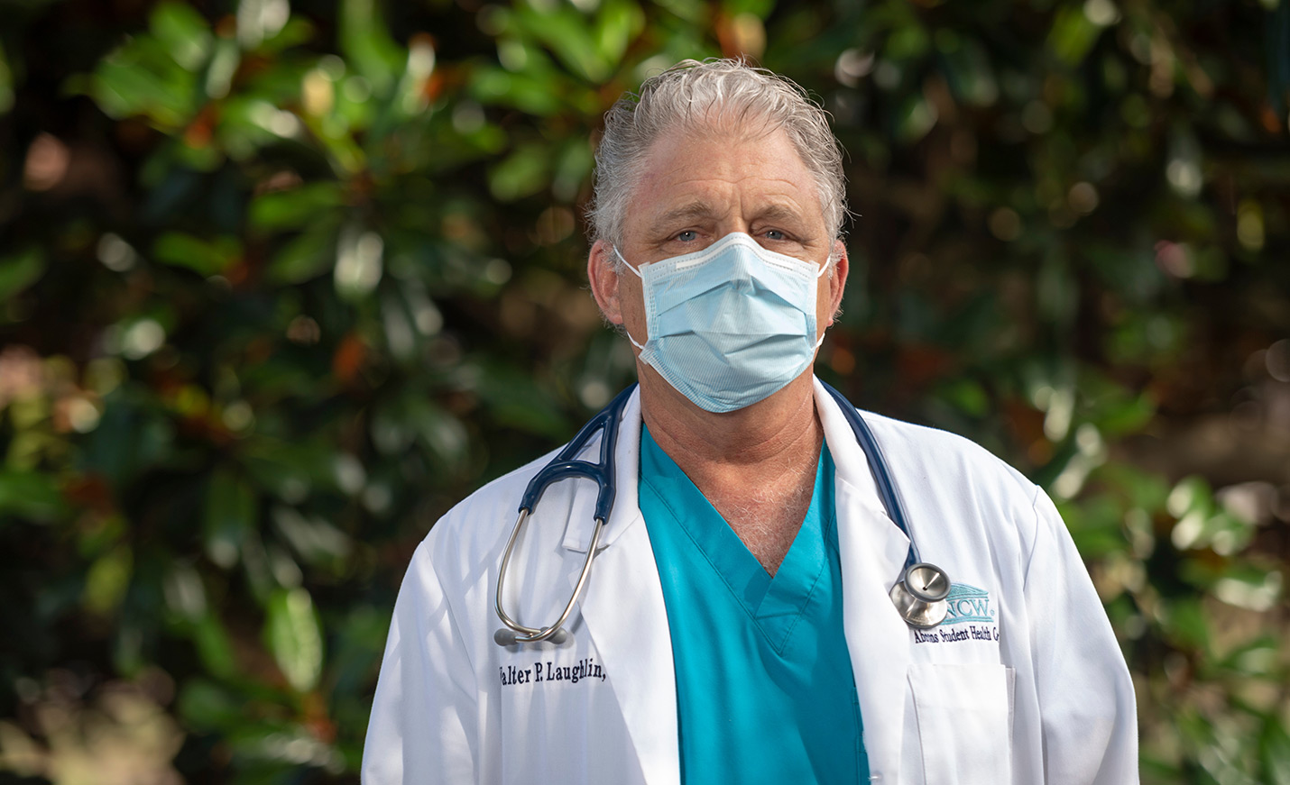 A man wearing a white medical jacket, scrubs and a paper mask. A stethoscope is around his neck.