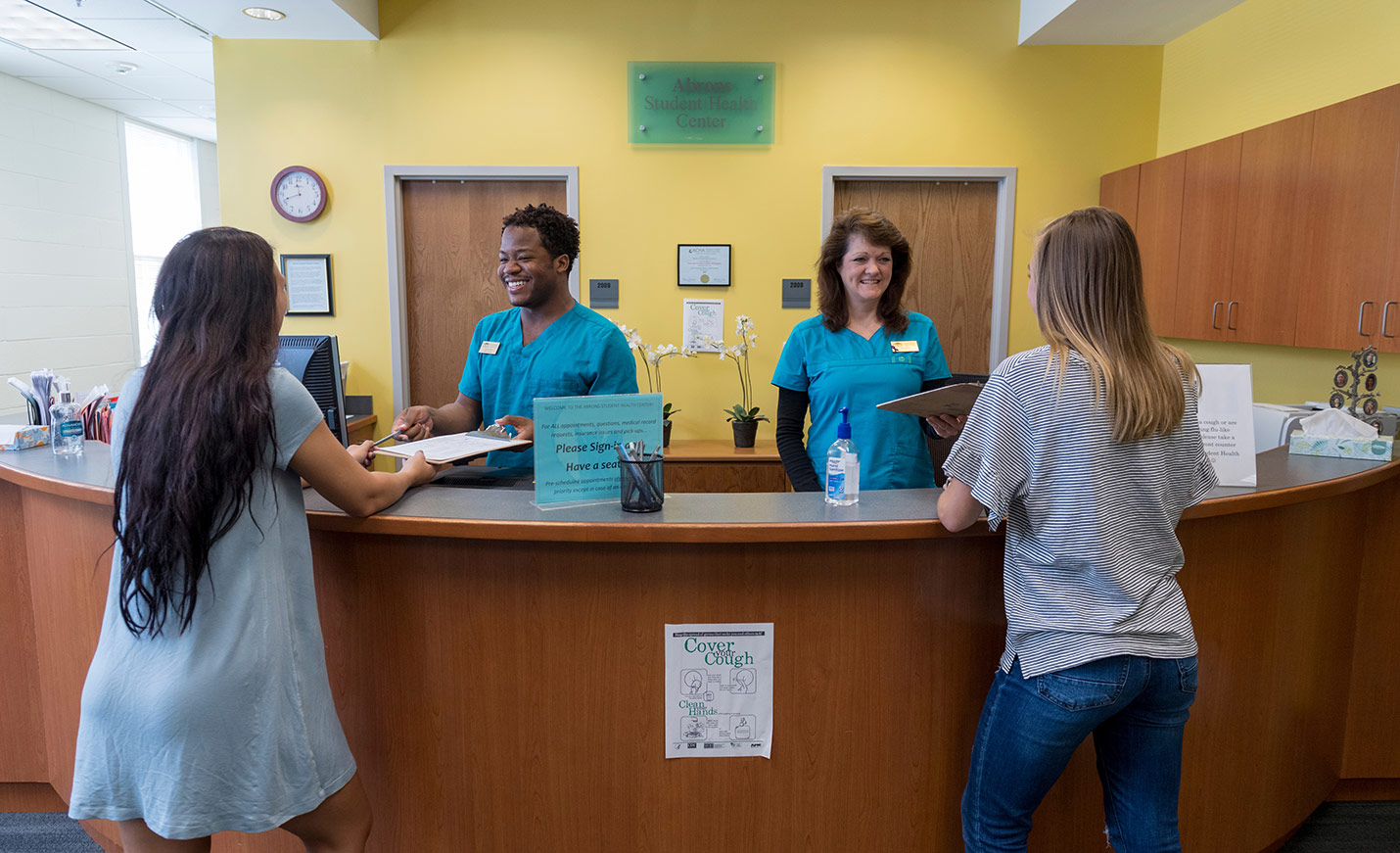 Two people in scrubs stand behind a medical office desk while two students with backs to camera talk with them