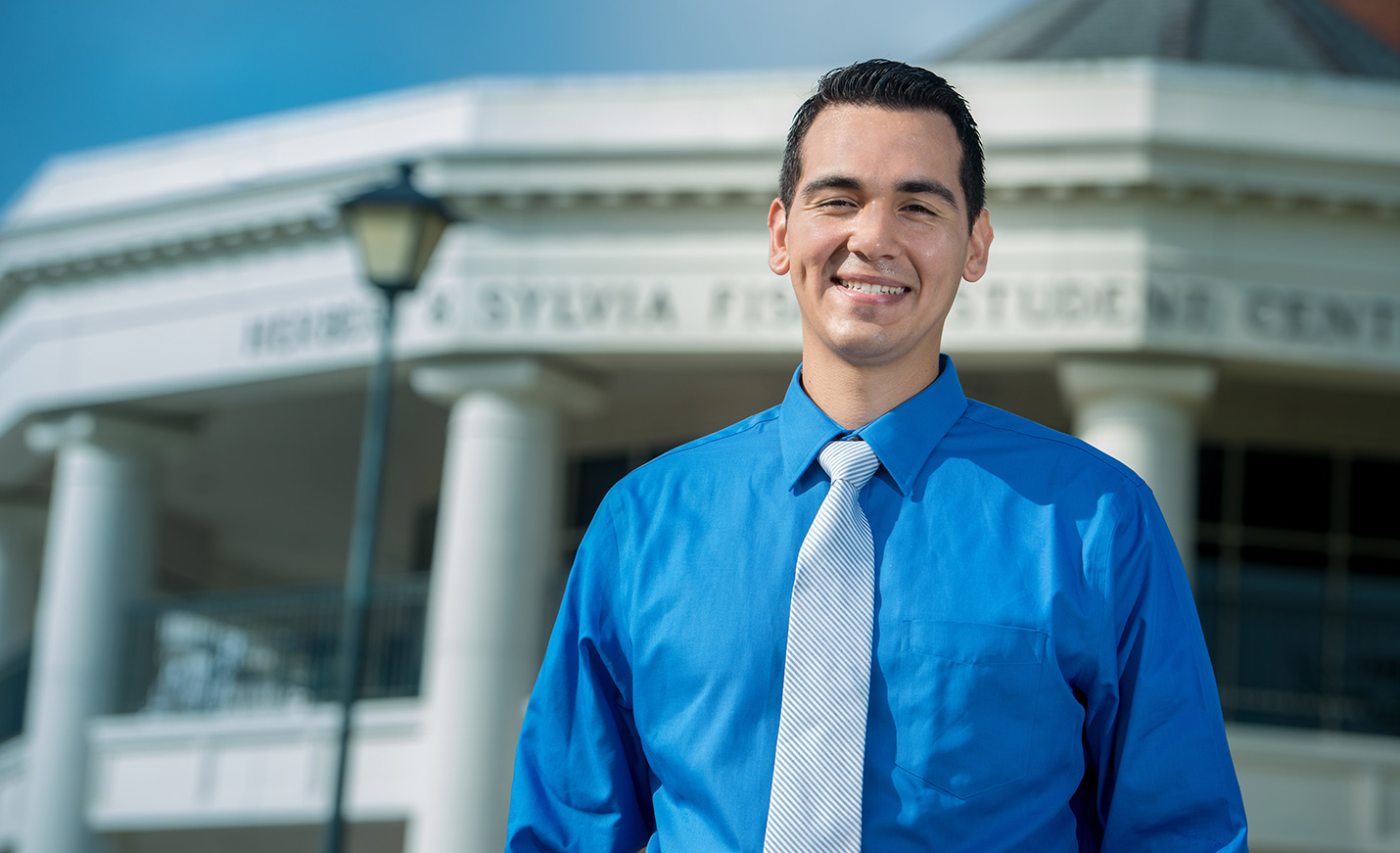 A man in a blue shirt and gray tie standing outside of the Fisher Student Center