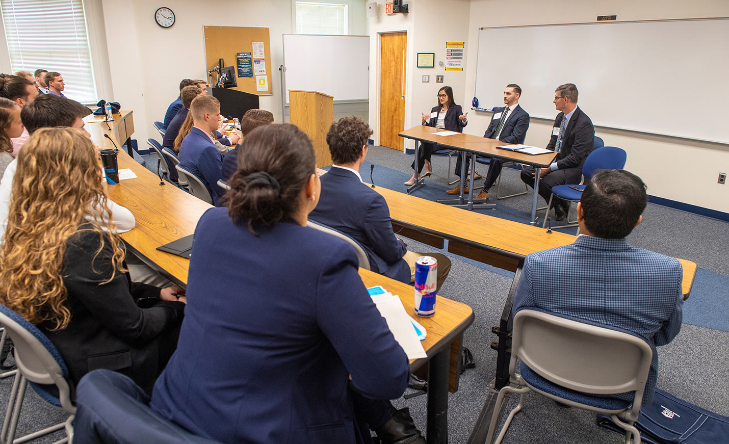 Three people talk to a class of students seated at tables in a lecture hall at UNCW