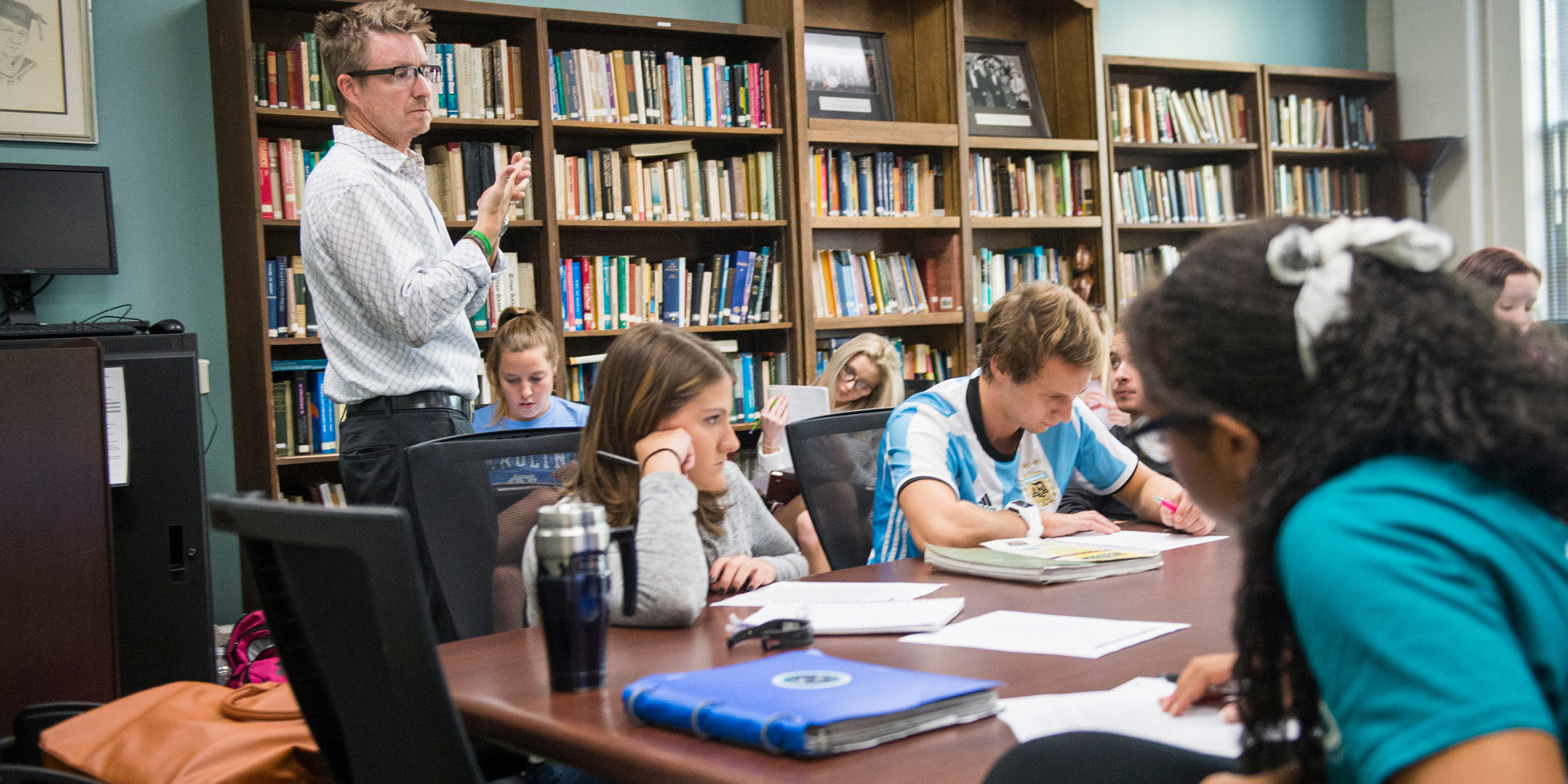 A professor gives a lesson to a room full of students with bookshelves lining the walls
