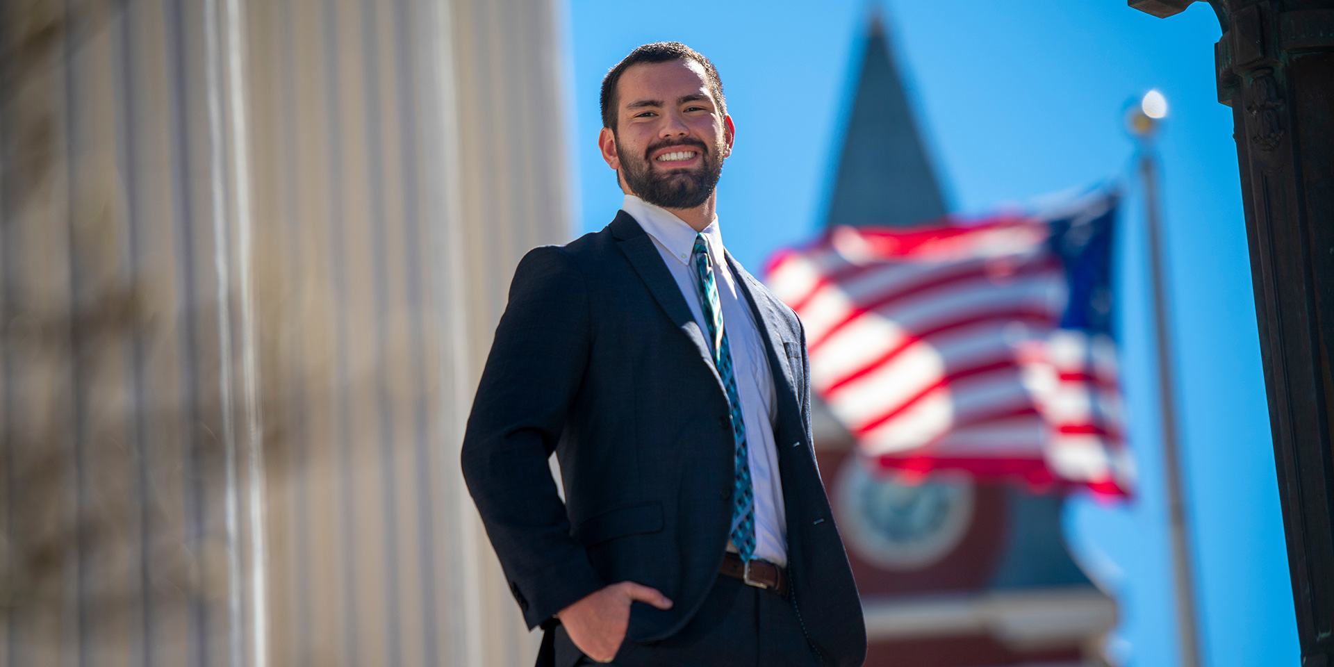 A student in a suit stands on City Hall’s steps with an American flag waving in the background