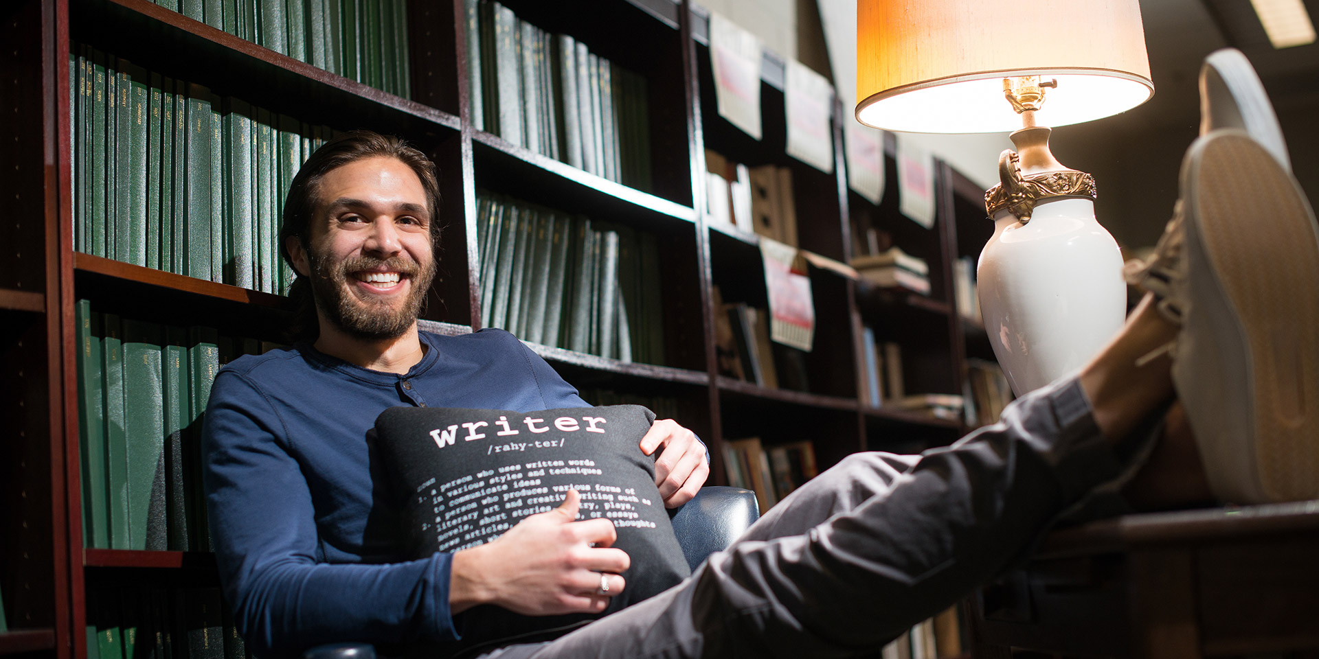 A person sits with their feet up, holding a pillow that defines the word “Writer”