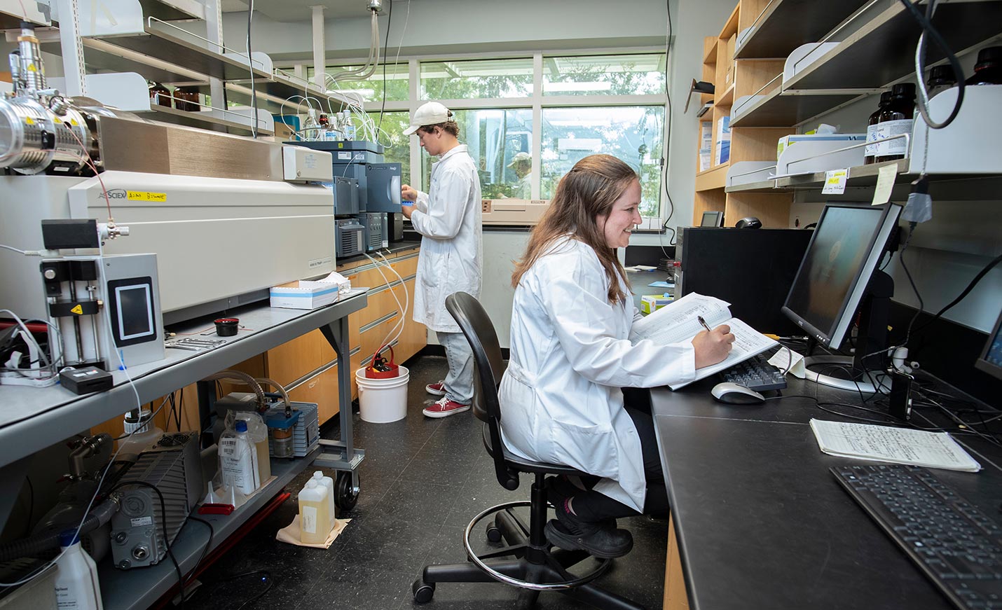 Two students working in a lab - one at a computer and the other, in the background, looking at a machine for results of a test