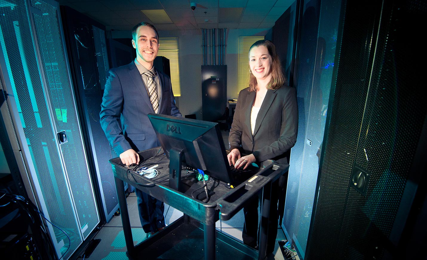 A woman stands in front of a computer sitting on a cart while a man stands next to her; both are smiling
