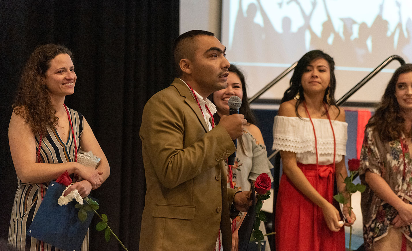 A man speaking into a handheld microphone while four women stand next to him and look on