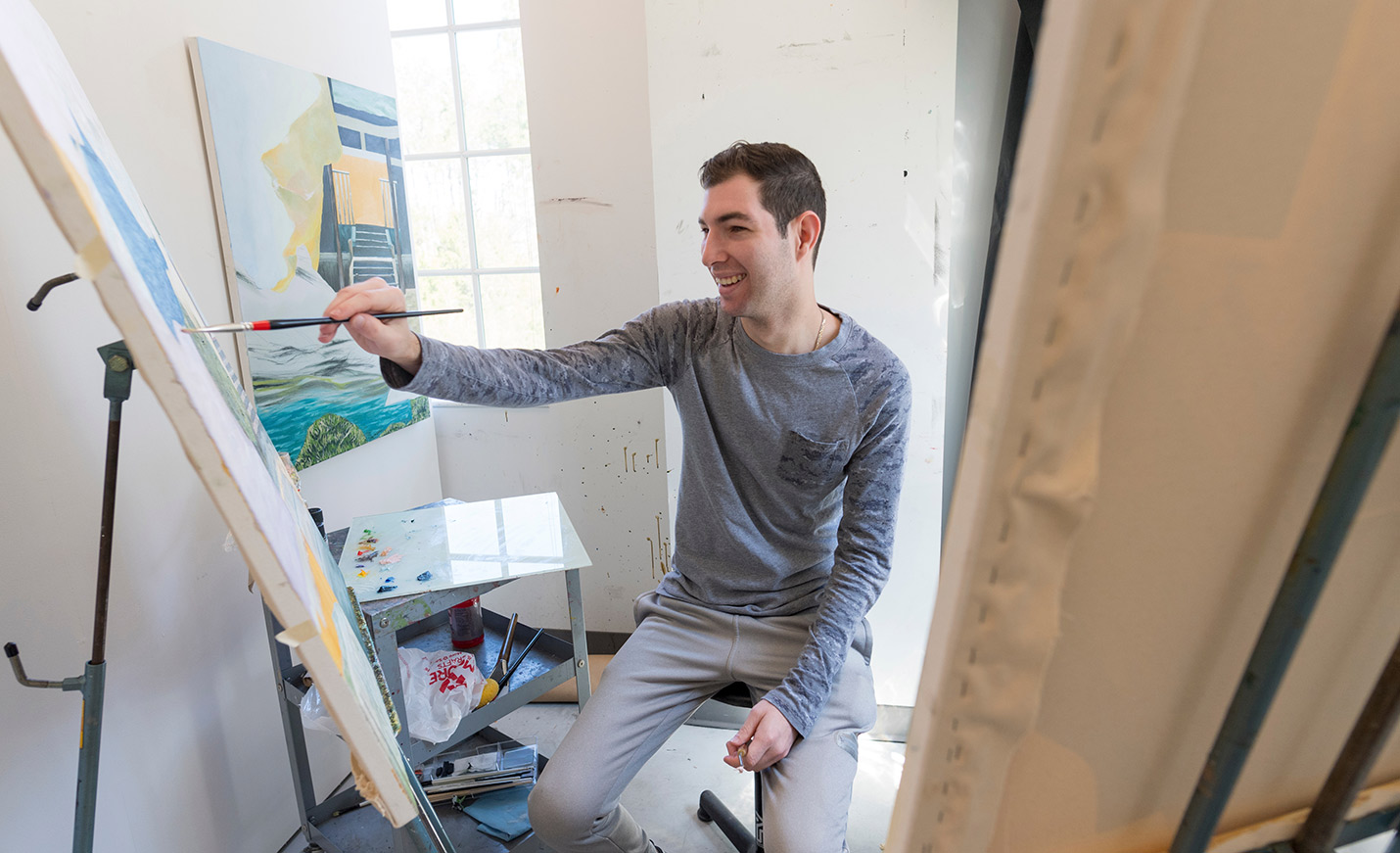 A student paints while seated in front of two canvases