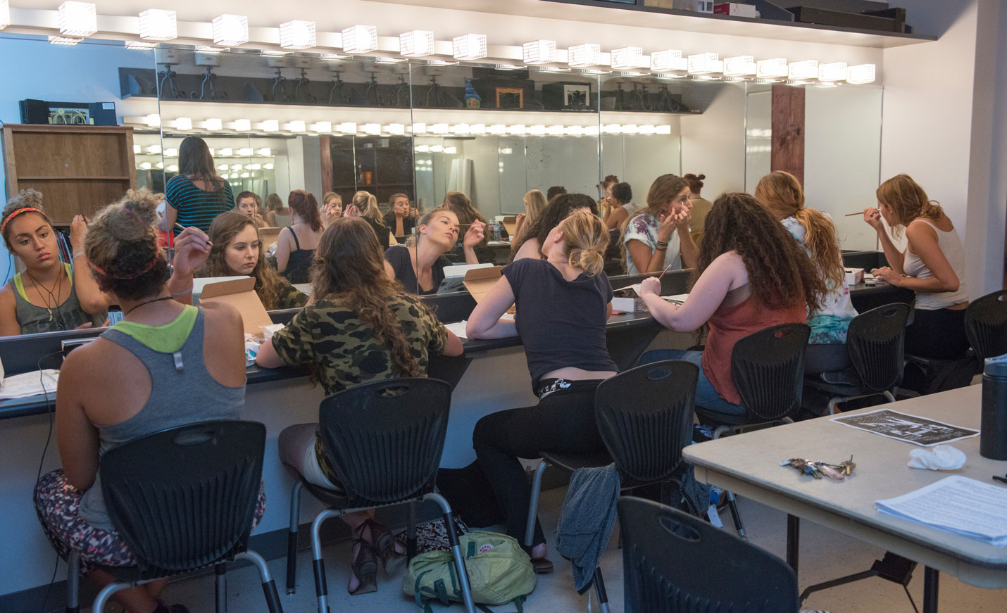 Theatre students applying makeup in while seated in front of a large mirror