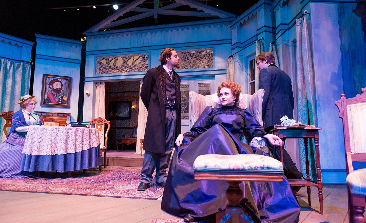 An actress in period dress stretched out on a chaise while two men stand near her and a woman in a cap is seated at a table nearby