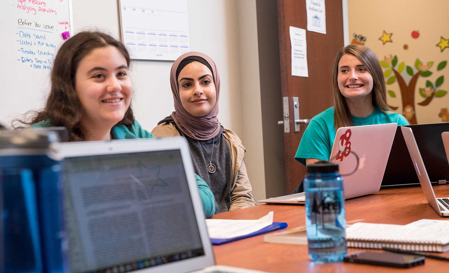 Three smiling students, one wearing a hijab, seated at a table with laptops open