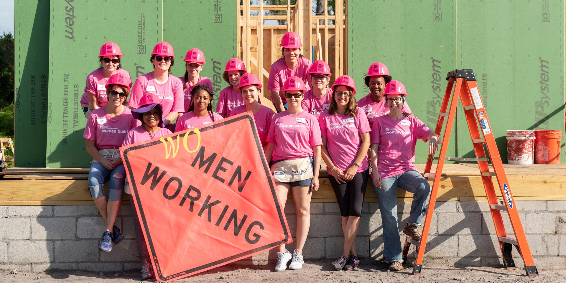 Group of people standing at construction site with construction sign that says “Wo” Men Working