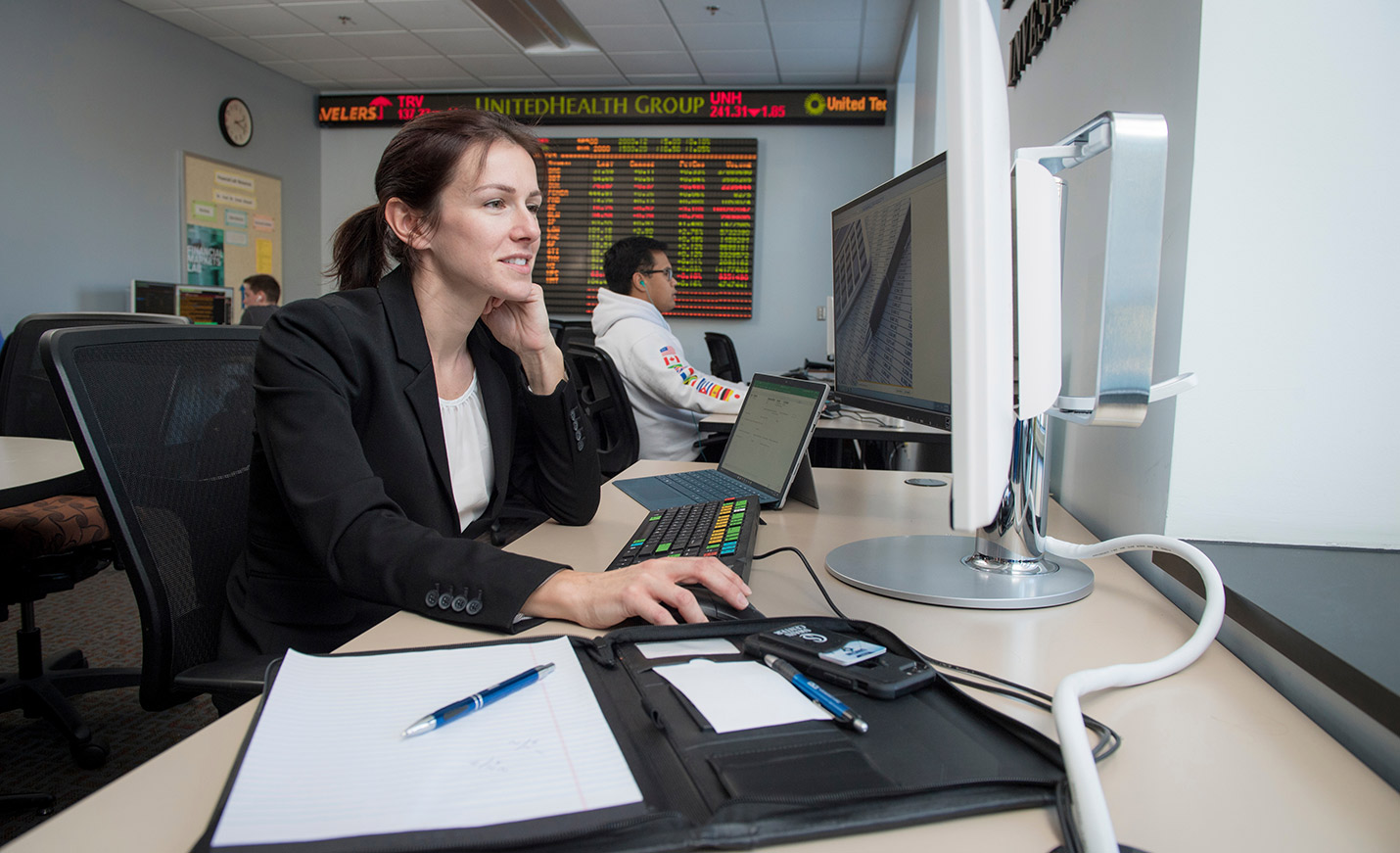 Two people, including one in the foreground, work on computers in a computer lap with the stocks ticker in the background
