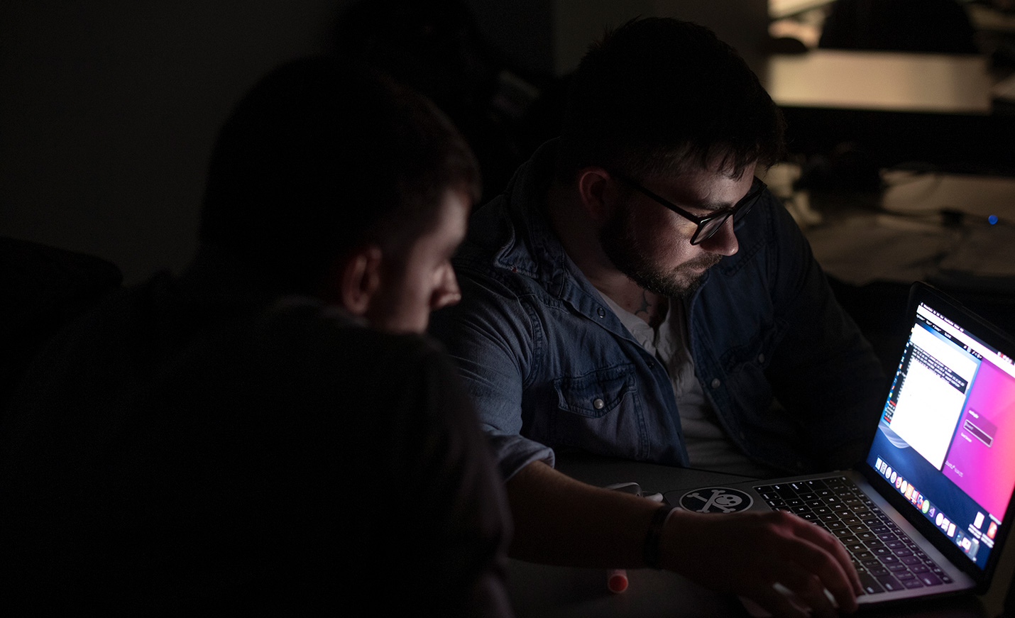 Two students work on a laptop in a darkened room