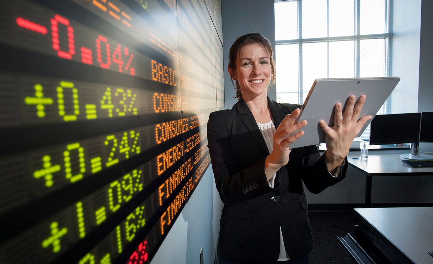 A woman holding a tablet stands in front of the stocks ticker in Congdon Hall
