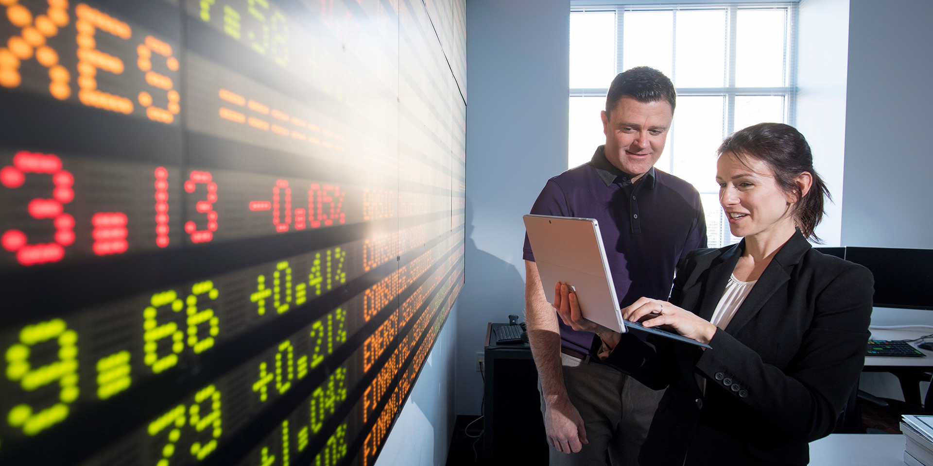 Two people discuss something on a tablet next to a large stock tickerboard