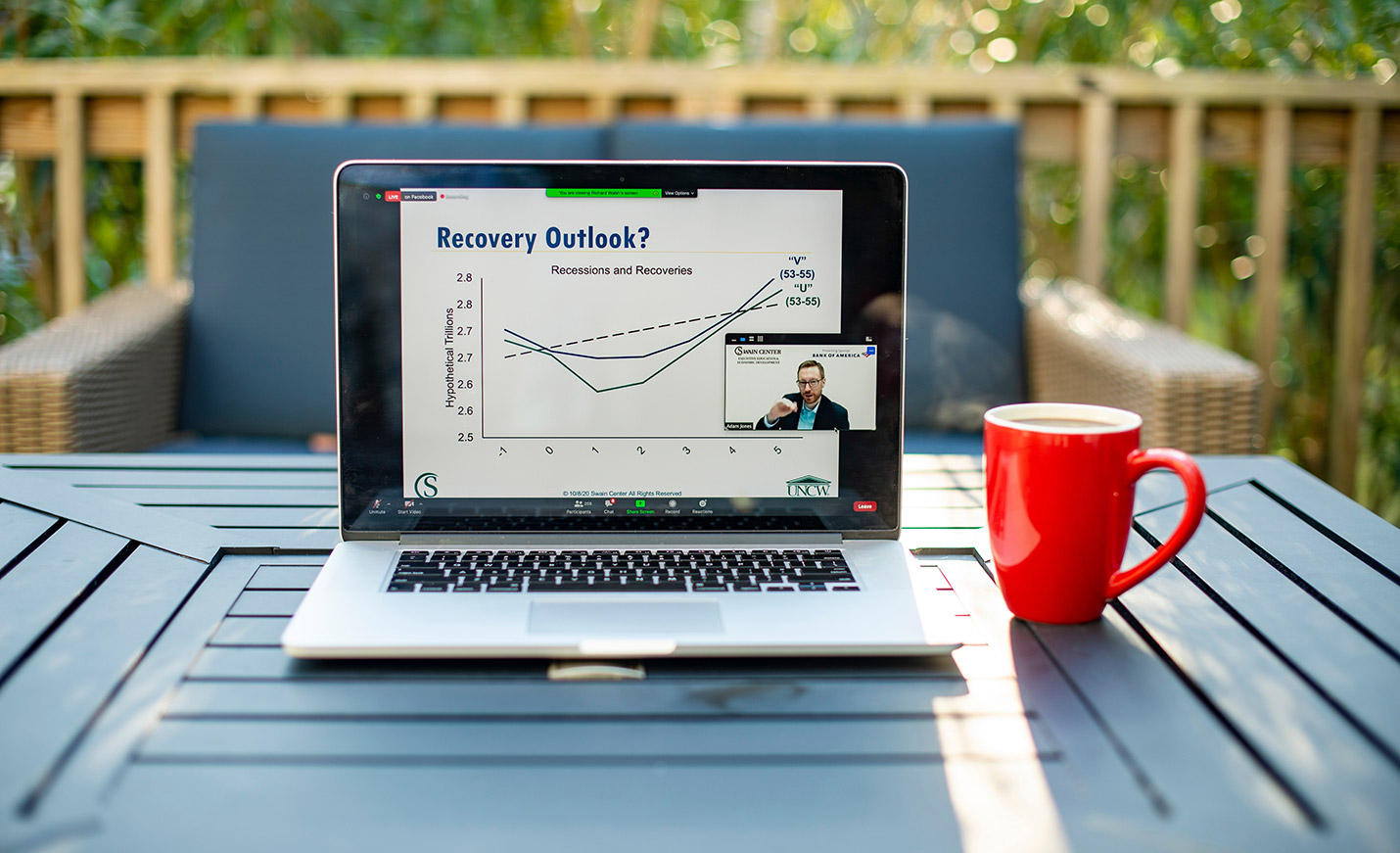 An open laptop on an outdoor table with a chart outlining economic information; a coffee cup sits next to it