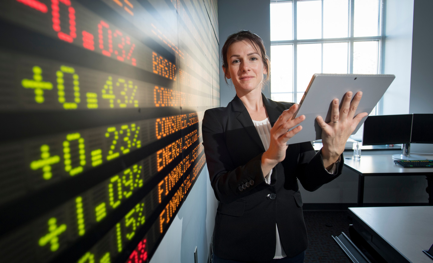 A woman holding a computer, standing in front of the stocks ticker in Congdon Hall