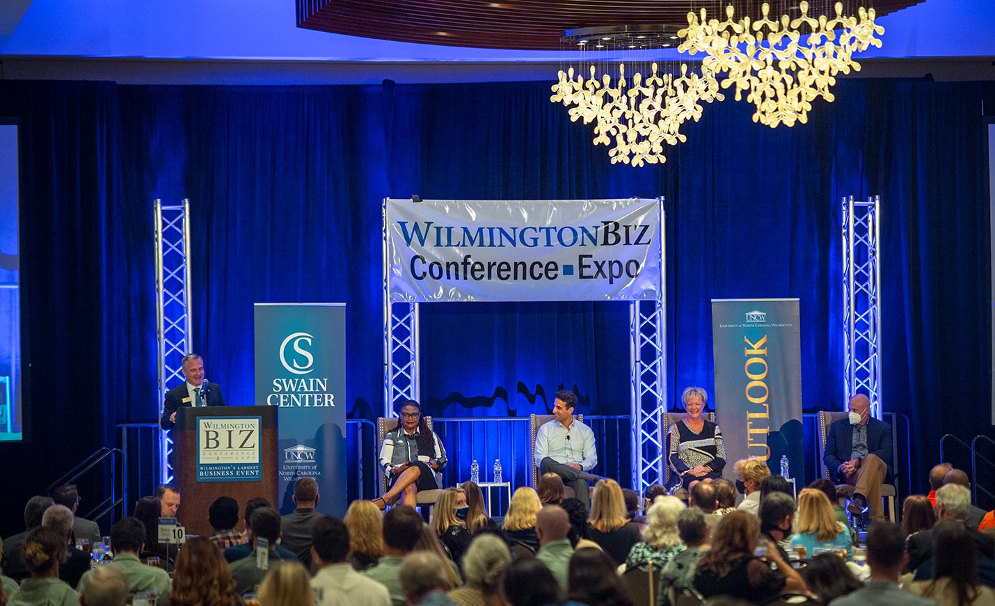 A moderator speaks at a podium with his head turned toward a seated panel at the Economic Outlook Conference