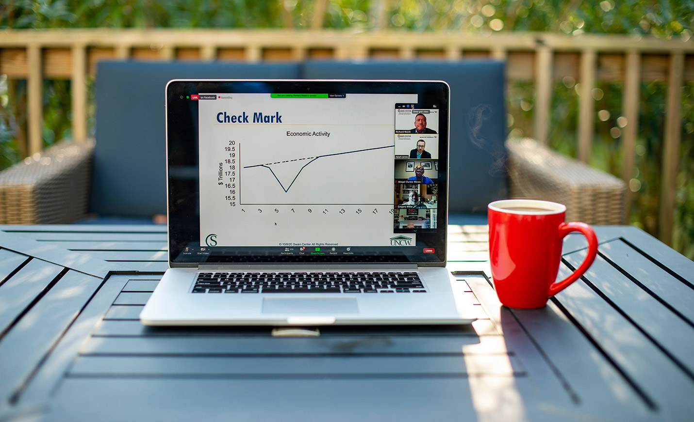 An open laptop on an outdoor table with screen displaying economic information; a coffee cup sits next to it