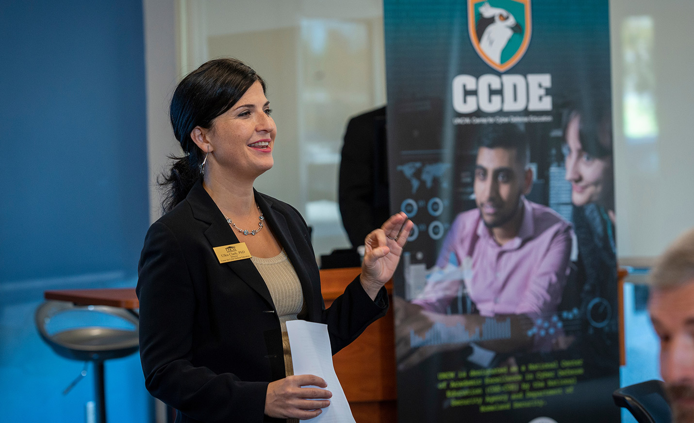 A faculty member speaks at an event with a screenshot in the background