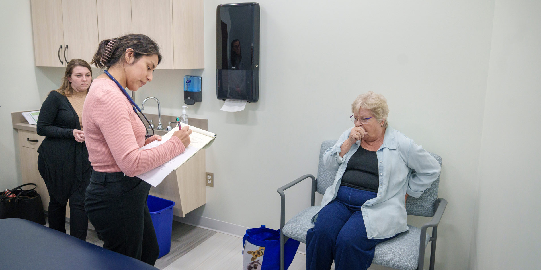 Three women in doctor's office. One, elderly, coughing, the other writing notes.