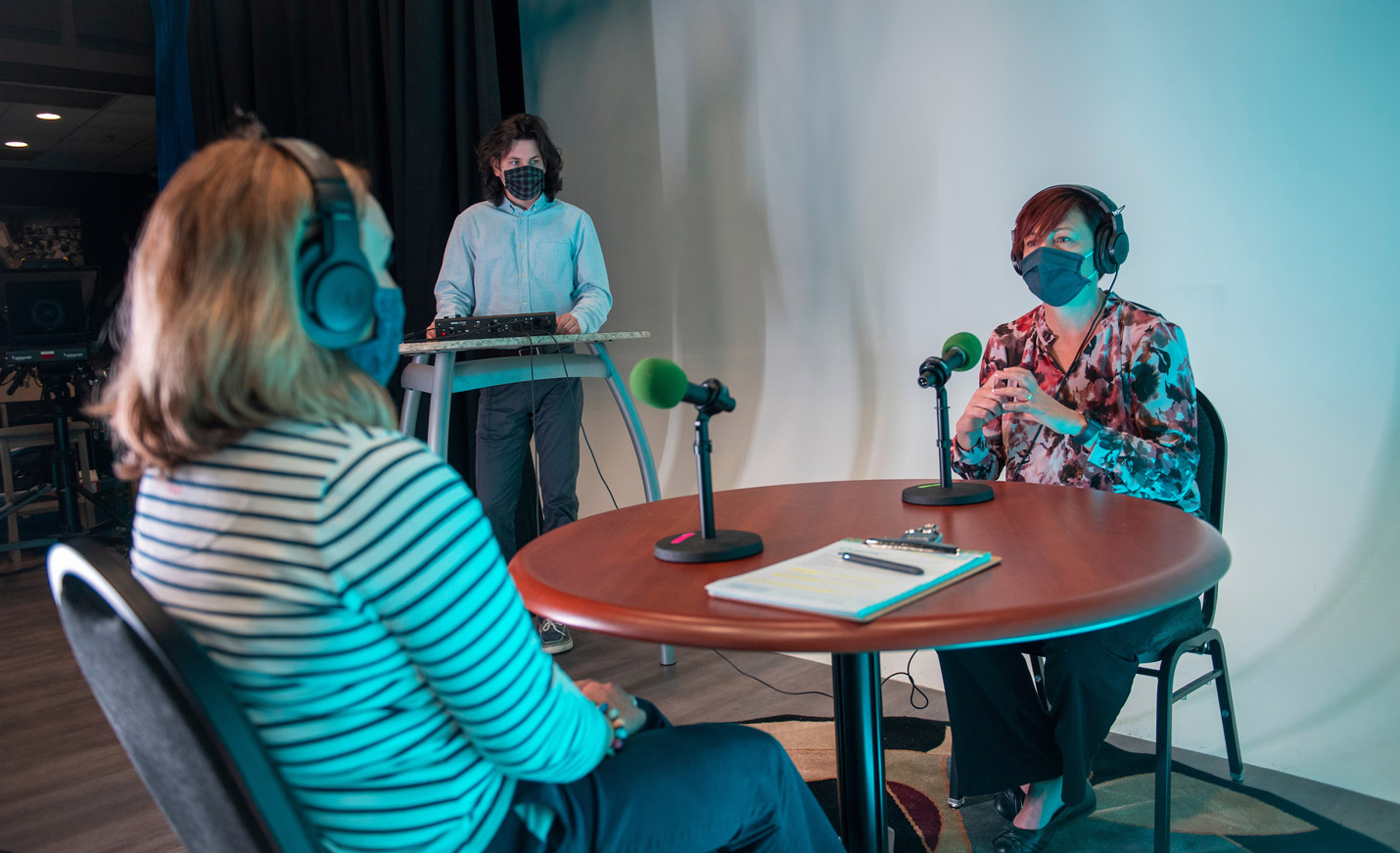 Two women wearing masks speak into a microphone for a podcast while a man stands at a production console