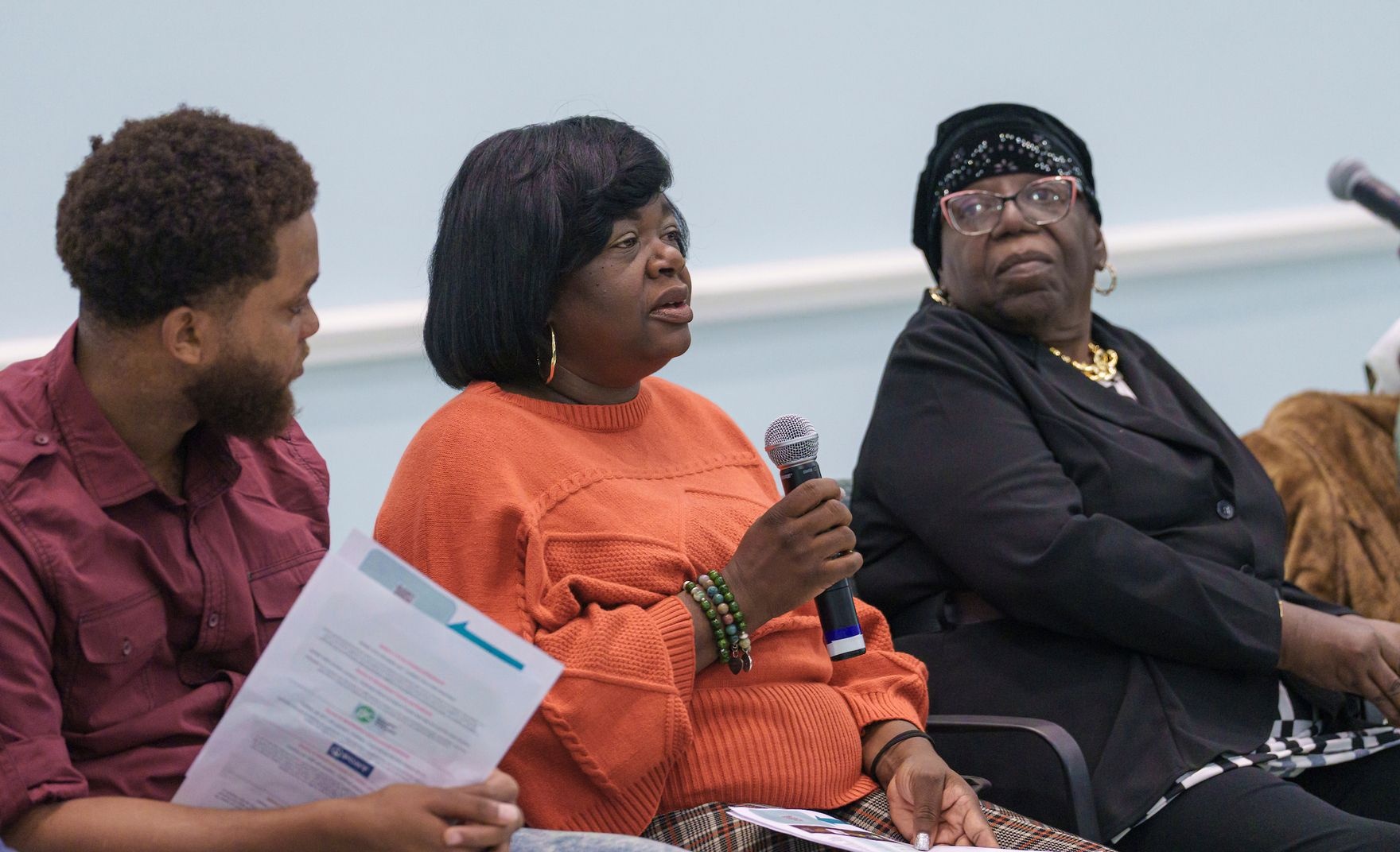 A seated woman holding a microphone speaks while a man on her right and a woman on her left look on