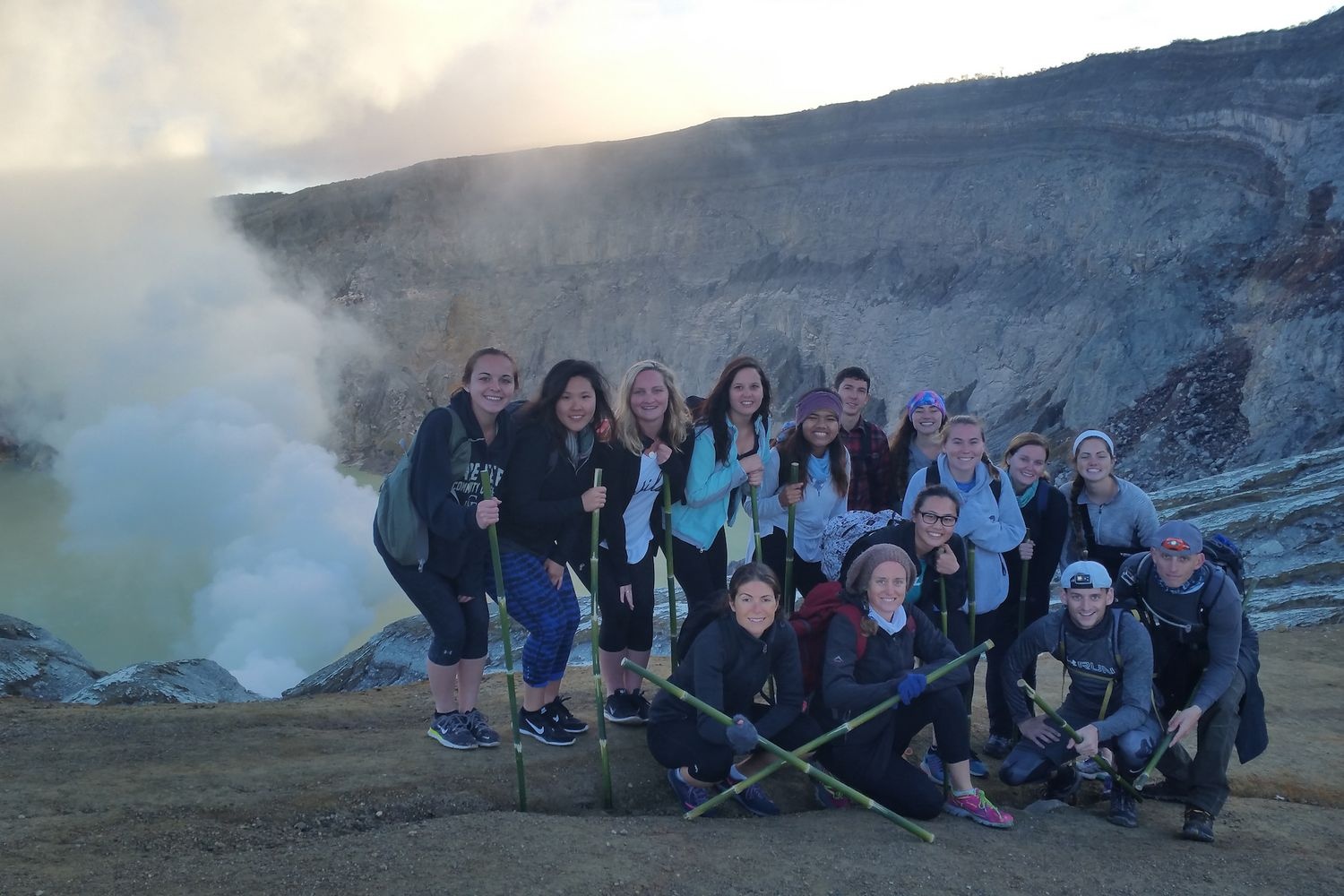 Group of hikers poses on a rocky overlook beside a steaming volcanic crater.