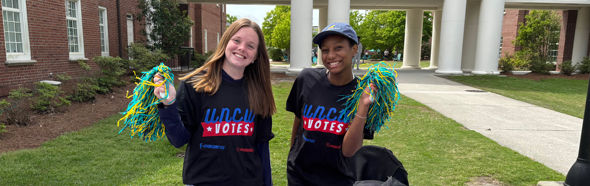Two students at a UNCW Votes booth outside of Fisher Student Union holding pom-poms