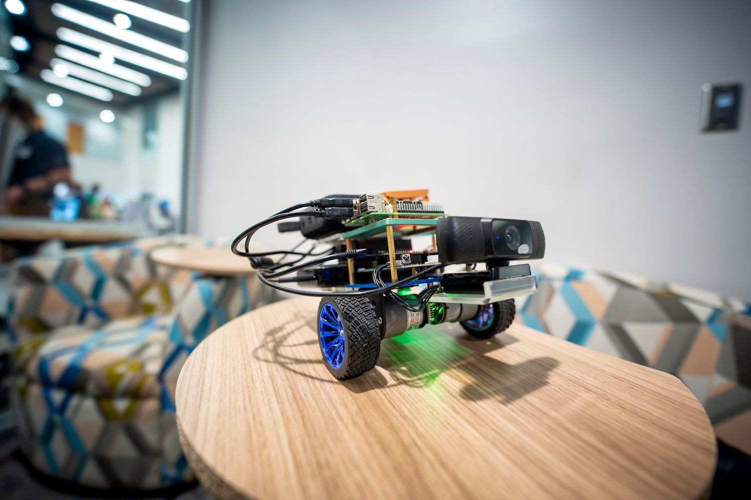 Small wheeled robot with blue rims, sensors, and circuit boards placed on a wooden round table in an indoor setting with patterned chairs and bright overhead lights.