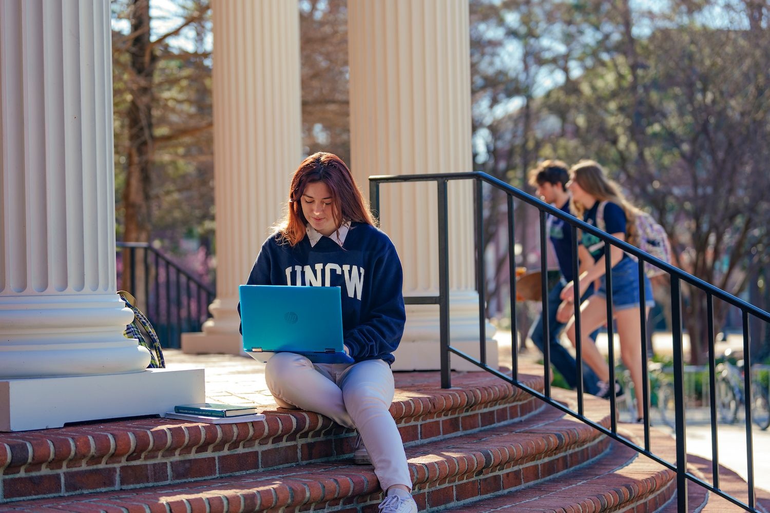 Student sits on campus steps using a laptop near white columns.