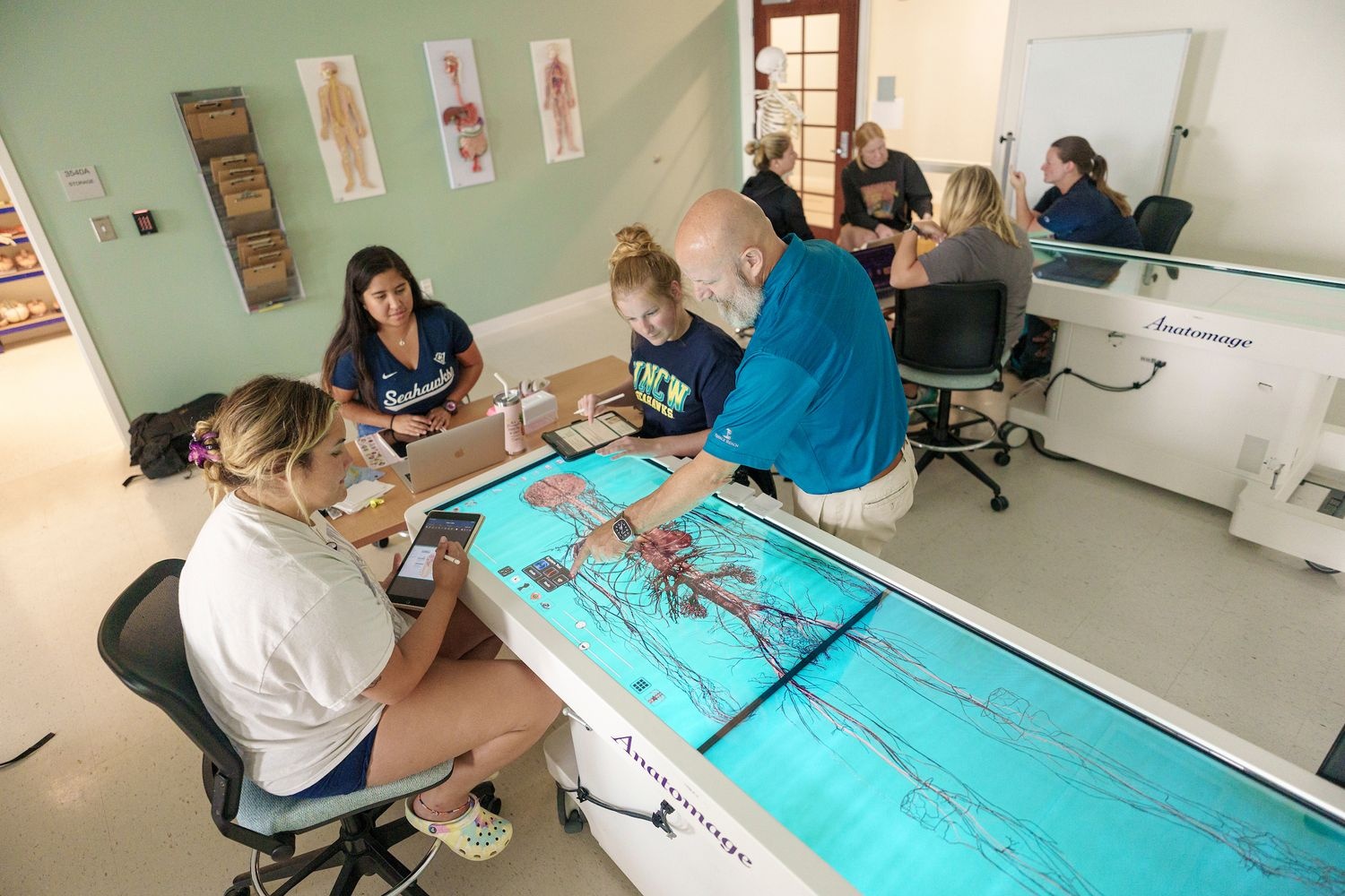 Students sit around a high tech anatomage table with their professor, learning about the digitally displayed (and life sized) human anatomy.