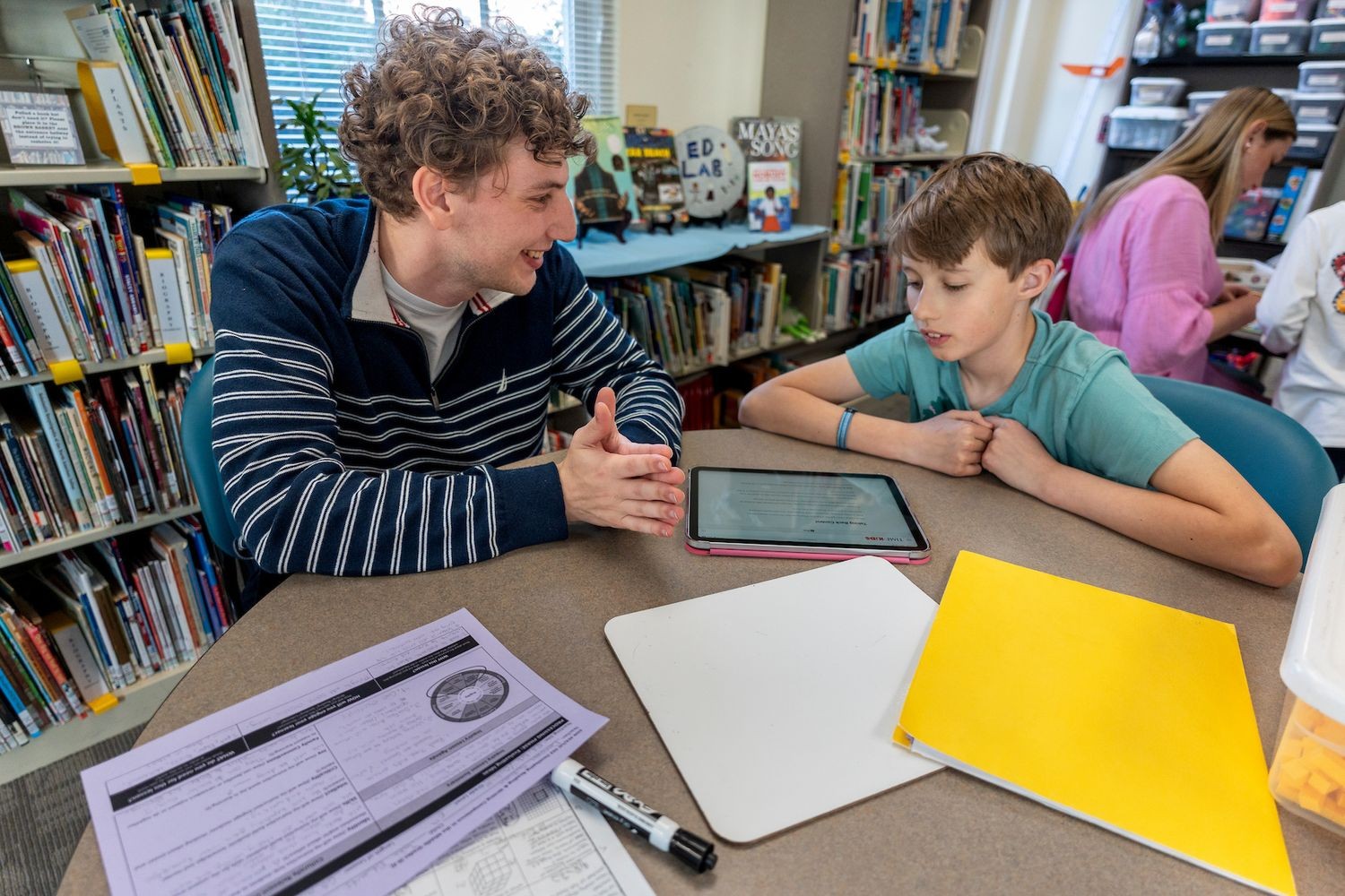 A student teacher works with a child on their assignment, in a room lined with children's book shelves.